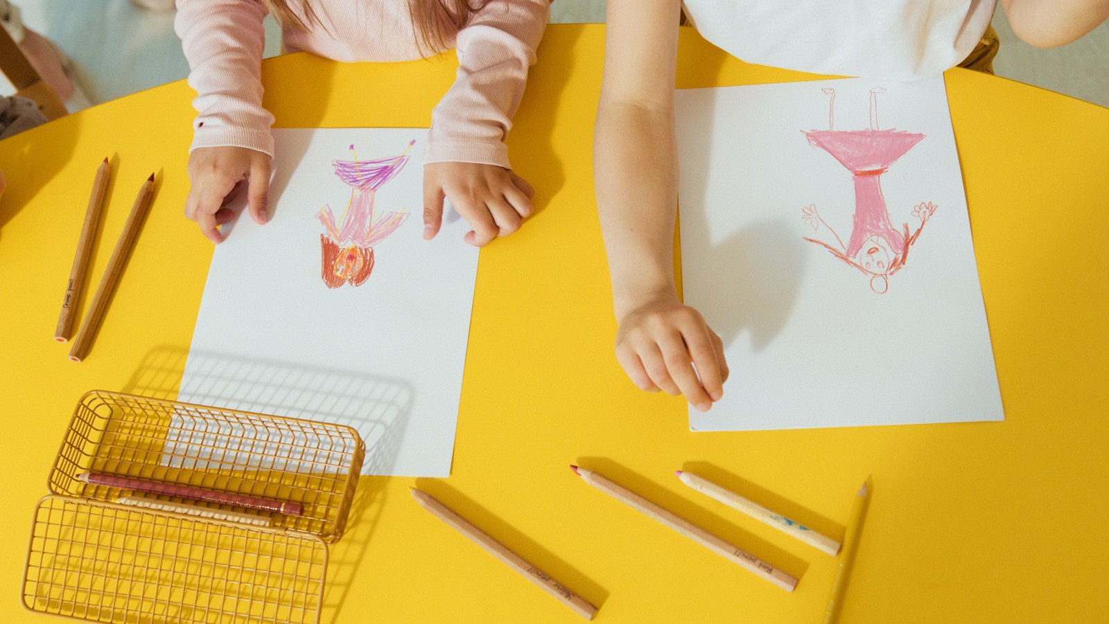 Two children drawing with colored pencils on paper at a yellow table