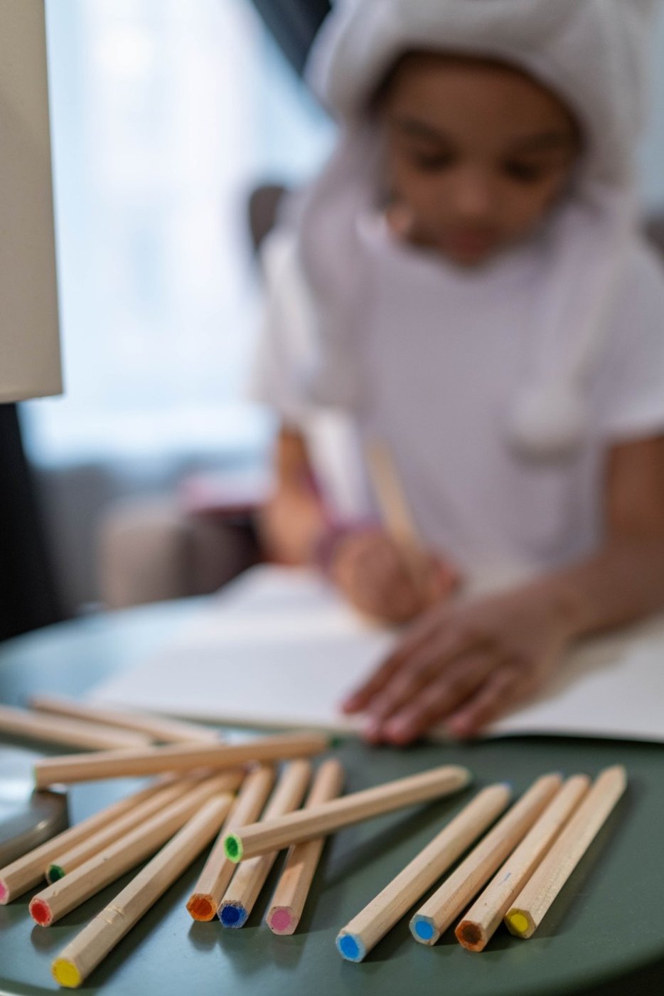 Colored pencils on a table with a child drawing in the background