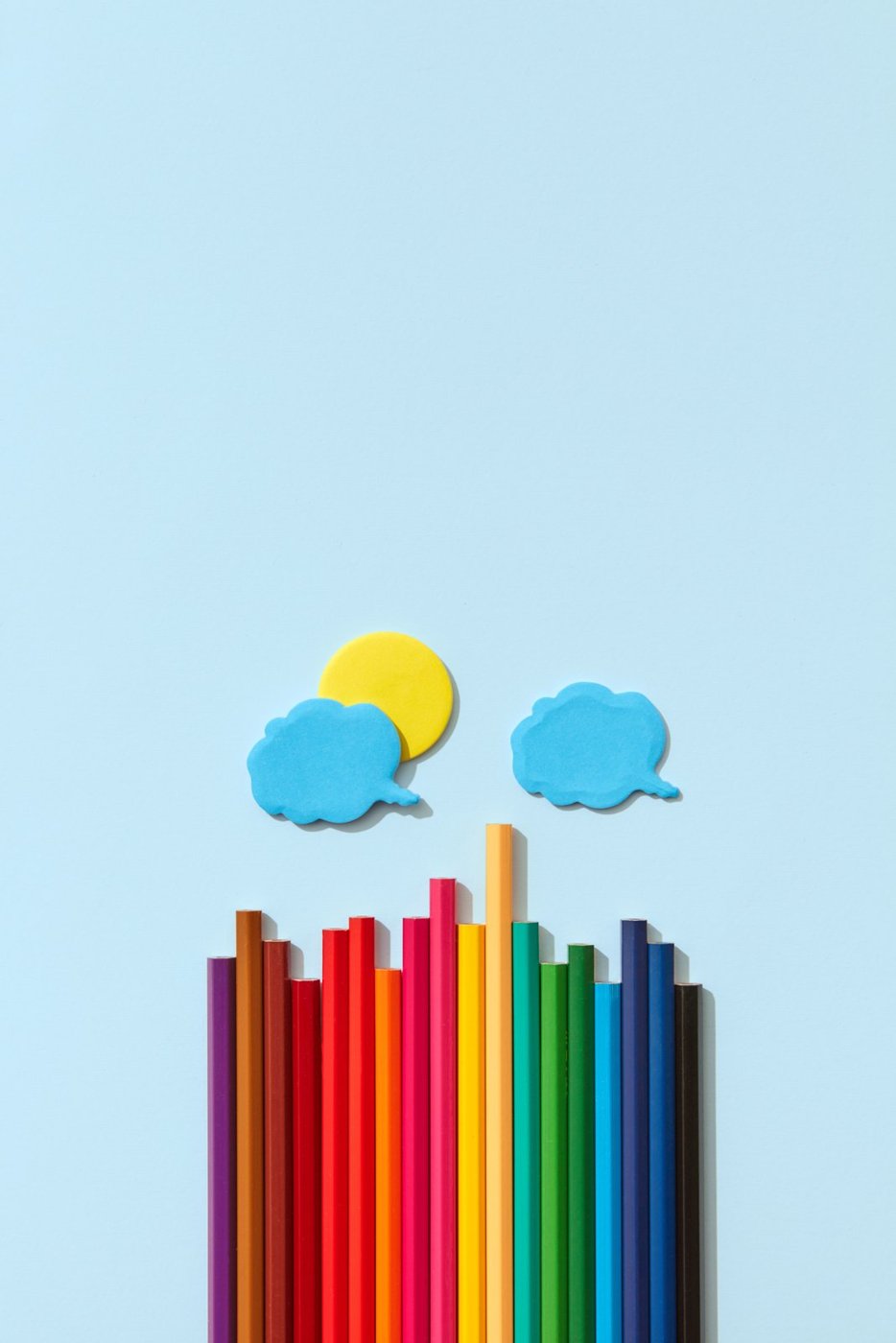 Colored pencils beside paper clouds and a sun on a blue background