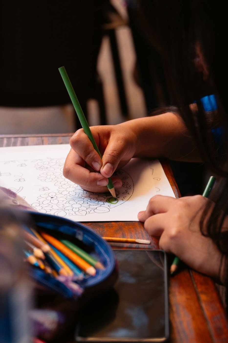 A child coloring on paper with a green pencil at a wooden table